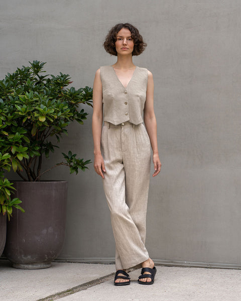 A light-skinned woman with short, curly brown hair stands centered against a textured light gray wall and a light gray paved ground. She is wearing a menique linen outfit consisting of a cropped, sleeveless vest in the color natural and high-waisted, wide-legged pants in the color natural. Black sandals are on her feet. Her posture is relaxed with arms at their sides.