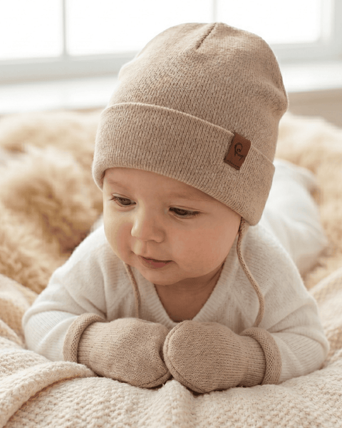A baby lying on a soft, textured cream blanket, looking gently downward. The baby is wearing a white long-sleeved top, a light beige knitted beanie with a small square brown logo tag, and matching beige knitted mittens.