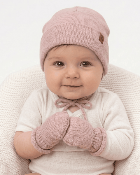 A softly smiling baby looking directly at the camera. The baby is wearing a white top, a dusty pink knitted beanie with a brown logo tag tied under the chin, and matching pink knitted mittens. The baby's mittened hands are held together over their chest.