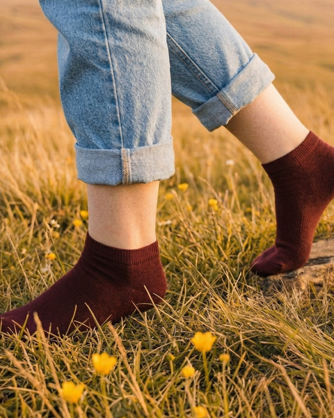 A close-up shot captures a person's lower legs mid-stride as they walk through a grassy field dotted with small wildflowers. They are wearing light blue cuffed denim jeans and deep burgundy ankle socks, bathed in warm golden hour light.