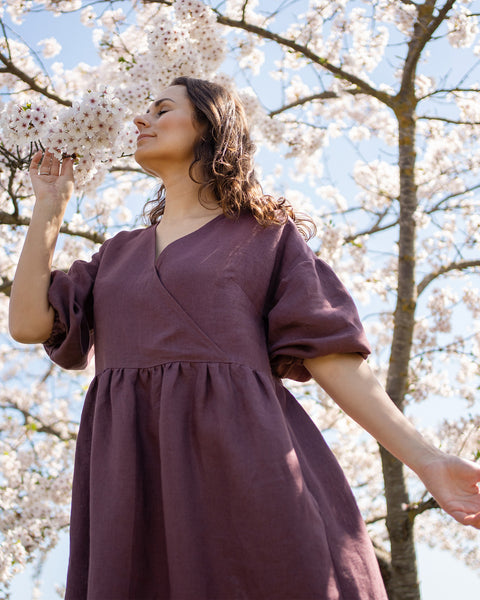 A person with dark, wavy hair and eyes closed stands outdoors, smelling the white blossoms of a tree. They are wearing a long, shadow purple V-neck dress with loose, elbow-length sleeves. The branches of the blooming tree are visible behind them against a bright blue sky.