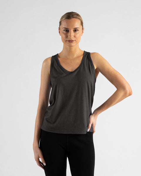 A front view of a blonde woman wearing a charcoal grey tank top and black leggings, standing with her left hand resting on her hip and her right arm relaxed at her side.