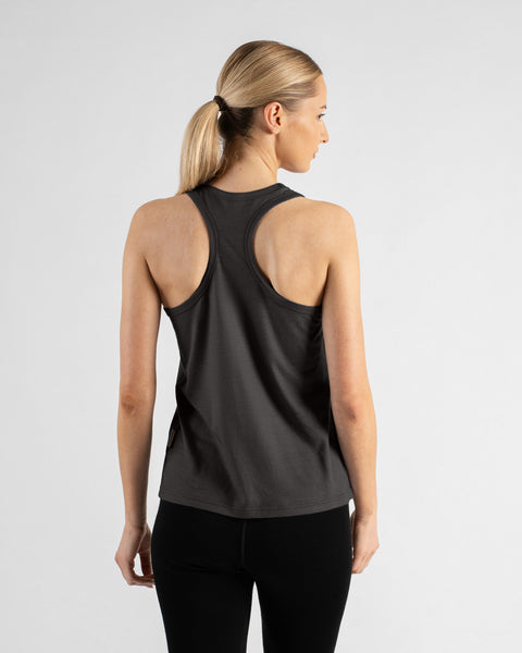 A back view of a blonde woman with her hair pulled into a low ponytail, wearing a charcoal grey racerback tank top and black leggings against a solid light grey background.