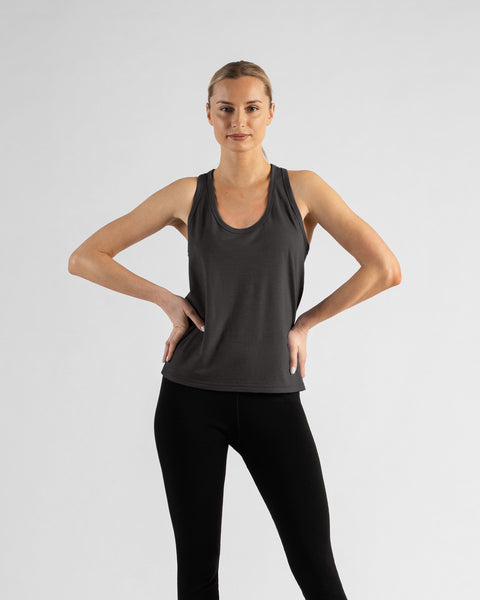 A front view of a blonde woman standing with both hands resting on her hips, wearing a charcoal grey scoop-neck tank top and black leggings against a light grey background.