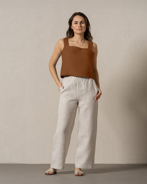 A woman with medium-toned skin and dark, shoulder-length hair stands facing forward against a plain, light-colored wall. She is wearing a teal, sleeveless top with a square neckline and linen menique pants with her one hand in the pocket.