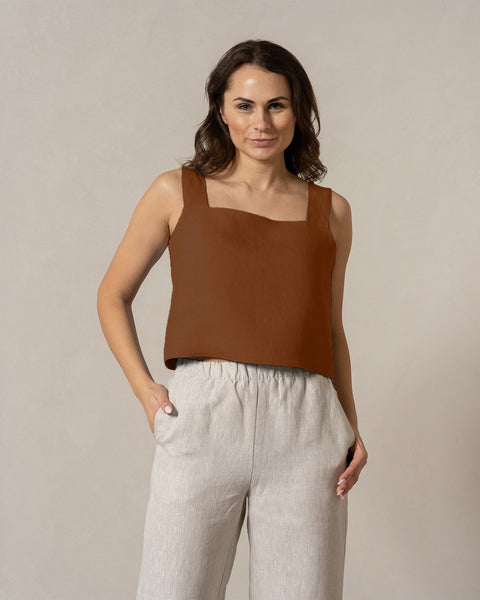 A woman with medium-toned skin and dark hair looks directly at the camera with a neutral expression. She is wearing a teal, sleeveless square-neck linen top and menique
linen pants, with her one hand in the pocket. The background is a plain, light beige color.