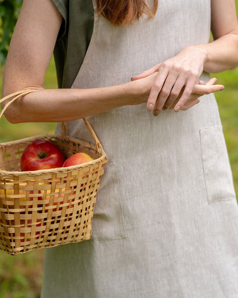 A close-up of a person wearing a natural-colored linen apron outdoors, holding a woven basket filled with apples against a green garden background.