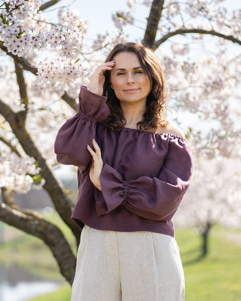 A woman with light skin and dark hair is shown from the waist up, smiling directly at the viewer. She is wearing a shadow purple, scoop-neck top. Her left hand, adorned with a ring on her ring finger, is casually raised to her chest, with her thumb touching her décolletage.