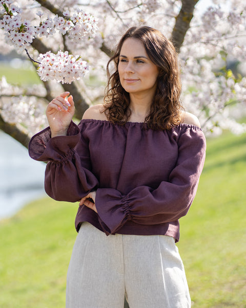 A woman with brown hair stands outdoors under a cherry blossom tree, looking to her right and smiling. She wears off-the-shoulder blouse with long, billowy sleeves gathered at the wrists and light-colored wide-leg pants. The background is a soft blur of green grass and more cherry blossom trees.