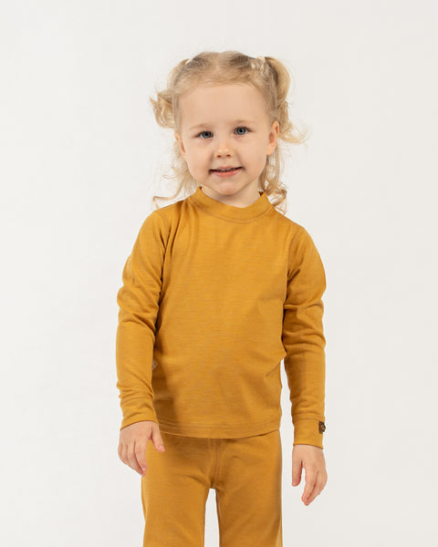 A young child with curly blonde pigtails, wearing a yellow long-sleeved top and matching pants, stands barefoot on a white background, looking directly at the camera with a gentle smile.