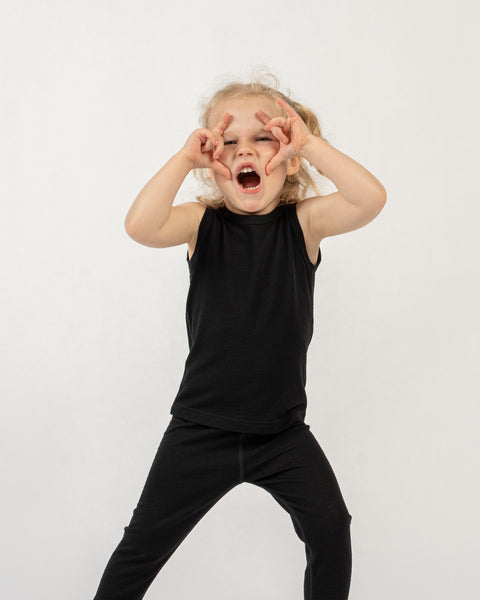 A young child with light, curly hair is standing against a plain white background. They are wearing a black sleeveless top and black leggings. The child is looking at the camera with a wide-open mouth, as if shouting or laughing, and both hands are raised to their face, with fingers forming circles around their eyes in a playful gesture.