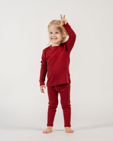 A young child with light curly hair stands facing forward, smiling broadly, and holding up both hands with two fingers extended on each, forming "peace" signs. The child is wearing a long-sleeved red top and matching red pants. The background is white.