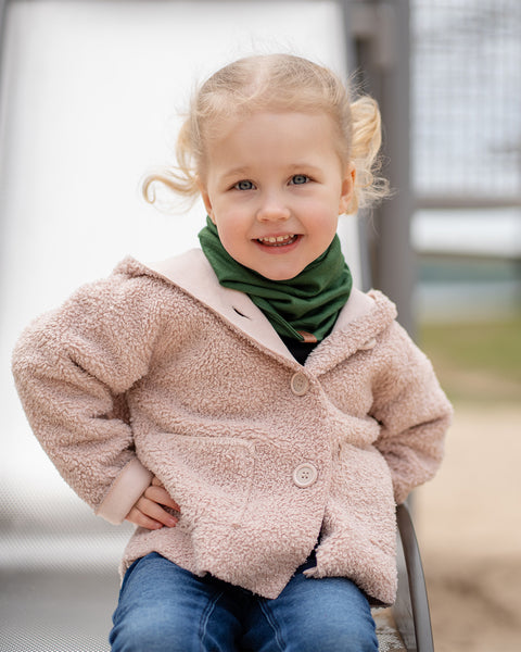 A young child with blonde hair and blue eyes smiles brightly while sitting on a slide, wearing a beige Menique jacket and a green neck warmer. The child's cheerful expression suggests a fun moment outdoors.