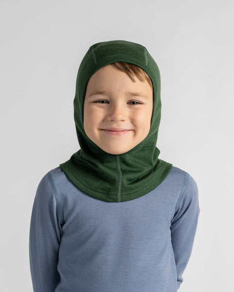 Portrait of a smiling young boy wearing a forest green knit balaclava hood over a dusty blue long-sleeve base layer.