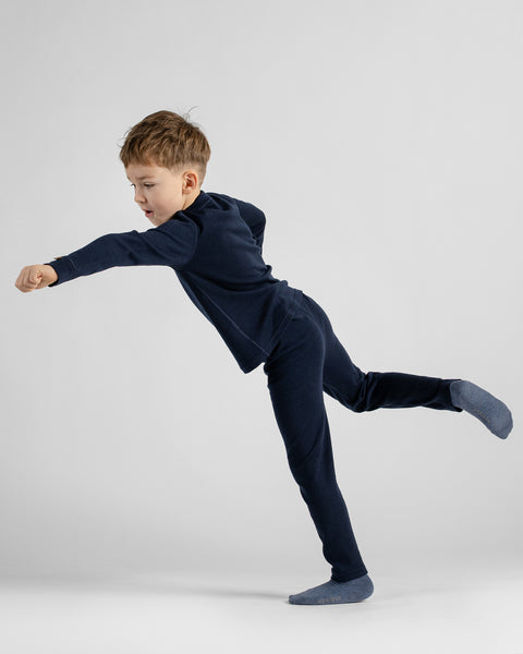 A young boy in a navy blue long-sleeve top and matching pants, wearing slate blue socks, captured mid-motion in a dynamic pose against a light gray studio background.