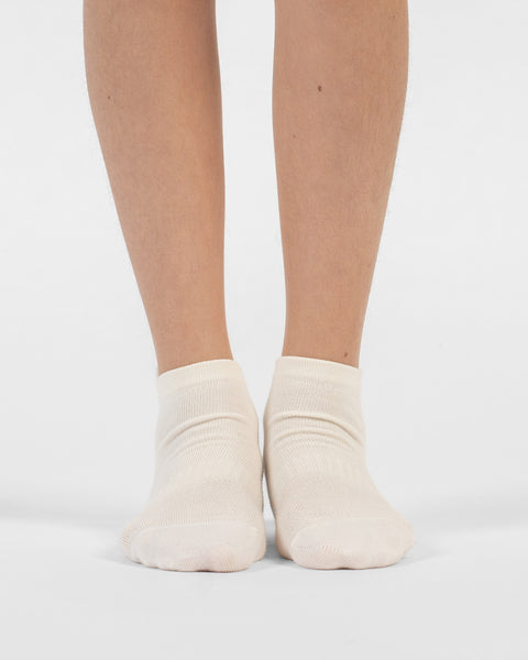 A front-facing view of a person's lower legs and feet standing flat on a white surface, wearing natural white cotton ankle socks.