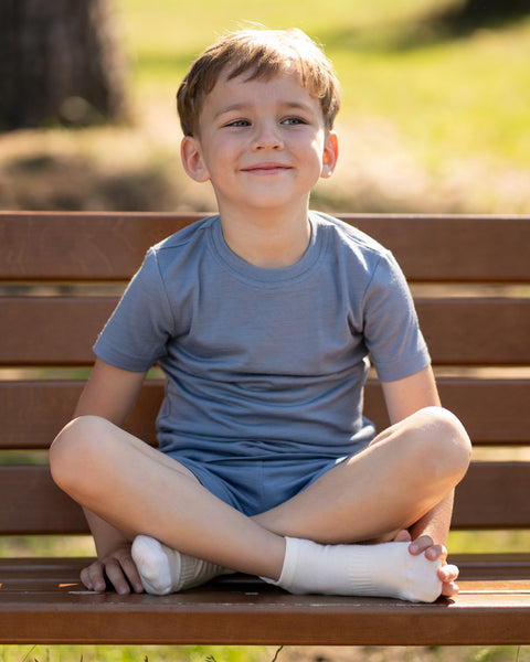 A young boy sitting cross-legged on a wooden park bench, smiling gently and looking off to the side. He is wearing a matching blue t-shirt and shorts set along with white ankle socks. The setting is a sunny, grassy park.