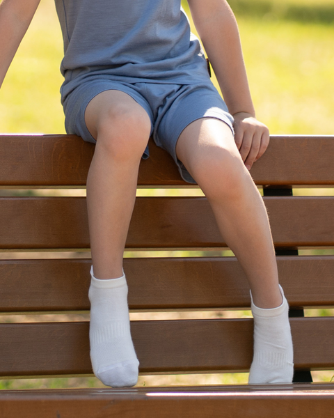 A child sitting on a wooden park bench with their legs dangling over the edge. They are wearing a blue short-sleeved top, matching blue shorts, and white ankle socks. The background is a softly blurred green outdoor setting.