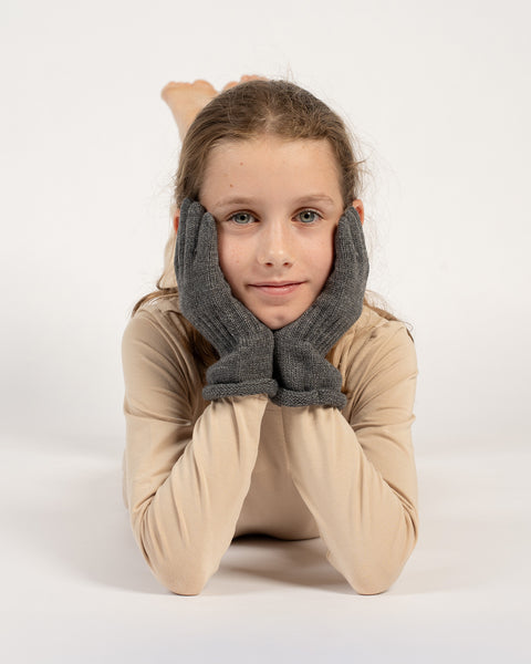 Child lying down with head resting on hands, wearing dark gray knitted cashmere gloves, beige outfit.