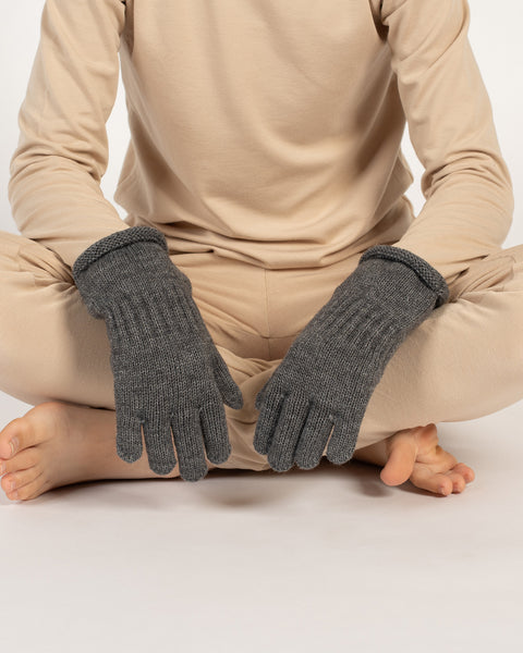 Child sitting cross-legged, wearing dark gray knitted cashmere gloves with beige loungewear.