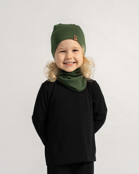 Smiling child wearing a dark green Merino wool beanie and matching neck gaiter, paired with a black outfit against a light background.