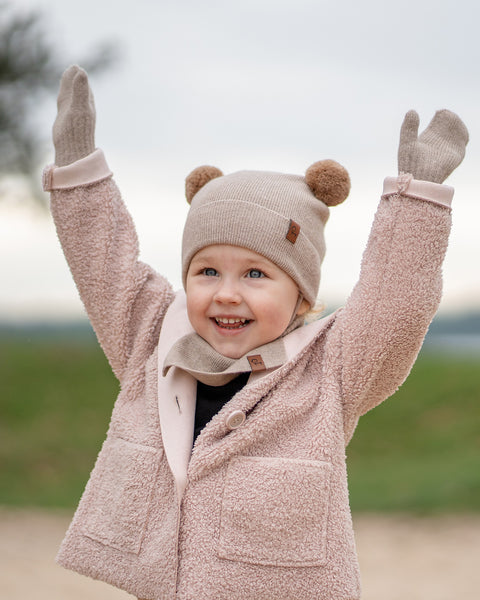 A joyous young child with blue eyes and an open-mouthed smile, stands outdoors with arms raised in excitement. They are wearing a beige knit hat with brown pom-pom ears, a matching beige scarf, beige mittens, and a light pink sherpa-style coat. The background is blurred with hints of green and brown.