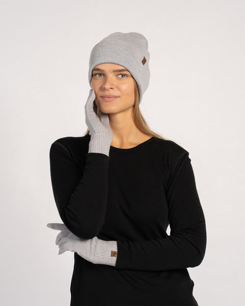 Woman wearing a light gray knit beanie and matching gloves, dressed in a black long-sleeve top, posing with one gloved hand touching her face and the other arm crossed against a plain background.