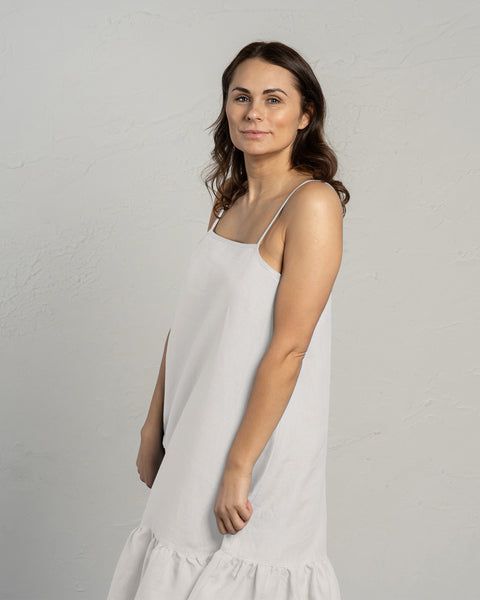 A woman with long, wavy dark hair and blue eyes looks directly at the camera. She is wearing a pure white linen dress with thin straps and a ruffled hem. Her right arm is slightly bent with her hand gently resting near her waist, while her left arm hangs loosely at her side. She is positioned against a textured light grey wall. The lighting is soft and diffused, casting minimal shadows.