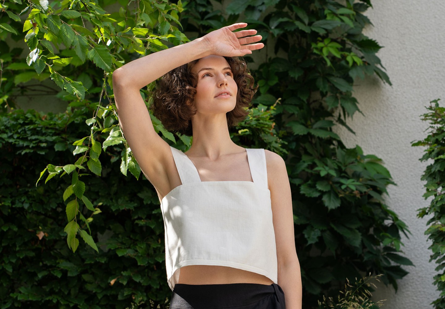 A woman wearing a sleeveless white linen crop top from menique shields her eyes from the sun while standing outdoors against a lush green background.