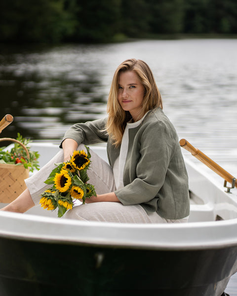 A woman sits peacefully in a white rowboat gently floating on a calm lake, holding a vibrant bouquet of sunflowers. She is dressed in an open-front stone green linen jacket layered over a pure white top, paired with relaxed natural linen pants.