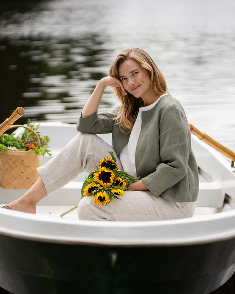 A smiling woman relaxes in a white rowboat on the water, resting her cheek gently against her hand. She is wearing a collarless stone green linen jacket, a white top, and natural linen pants, with a bunch of bright sunflowers resting on her lap.