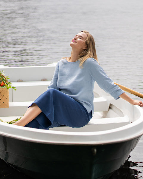 A woman relaxes inside a small white rowboat floating on a calm lake, leaning back with her face tilted toward the sun and eyes closed. She is wearing a long-sleeve, cloudy blue linen top paired with wide-leg, storm blue linen pants.