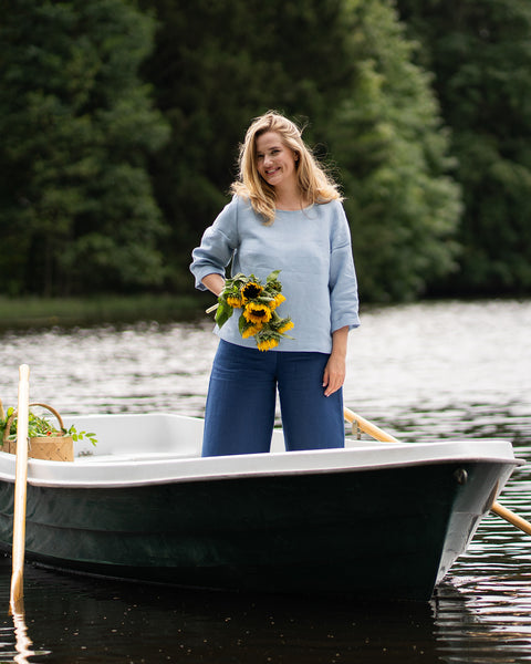 A smiling woman stands confidently in a white rowboat on a lake, holding a vibrant bouquet of yellow sunflowers. She is casually dressed in a long-sleeve, cloudy blue linen top and wide-leg, storm blue linen trousers.