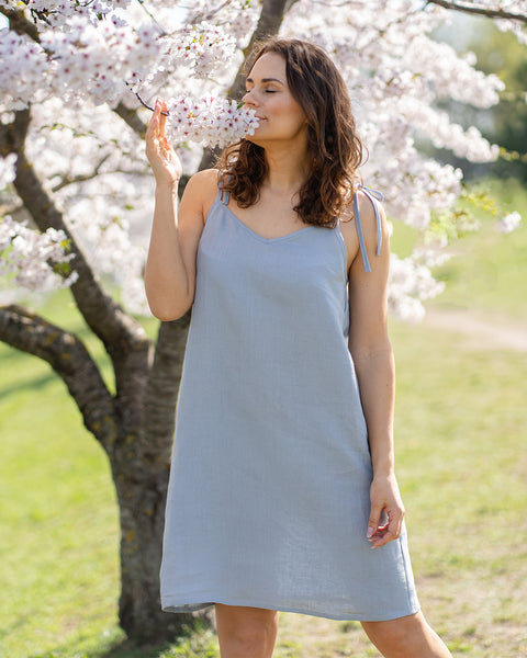 A woman wearing a cloudy blue linen slip dress stands beside a tree covered in white blossoms and gently smells a cluster of flowers.