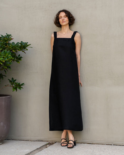 A fair-skinned woman with short, curly dark hair stands against a textured light brown wall. She is wearing a sleeveless, maxi linen dress with a square neckline and black sandals. To her left is a large potted green plant.