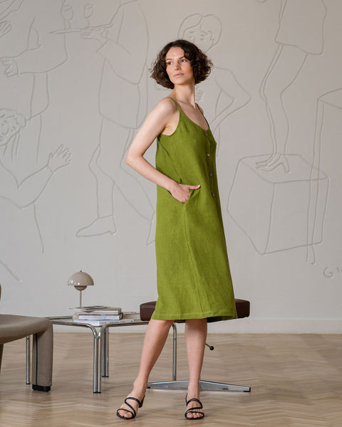 A woman with short, curly brown hair, wearing a sleeveless, button-front, forest green midi-dress with pockets, stands with one hand in a pocket. She wears black open-toed sandals. She is looking to her left. Behind her is a light-colored wall with white raised line art depicting abstract figures.