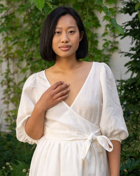 A person with short, dark hair and what appears to be Asian features is looking directly at the camera with a neutral expression. They are wearing a pure white wrap dress with a V-neck and elbow-length sleeves, and their right hand is gently placed on their chest. Lush green foliage is visible in the background.