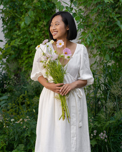 A smiling person with short, dark hair and what appears to be Asian features is holding a bouquet of light pink and white flowers. They are wearing a pure white wrap menique dress with elbow-length sleeves and are standing outdoors amidst lush green foliage and other flowers.