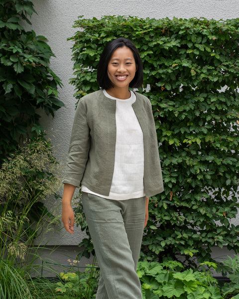A woman wearing a white top, gray jacket, and gray pants poses outdoors against a green leafy background.