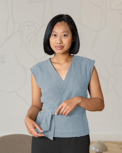 A woman with shoulder-length dark hair looks directly at the camera while wearing a menique cloudy blue, linen sleeveless top and pure black pants. Her hands are gently touching the tied section of the top at her waist. She stands indoors against a light gray wall with subtle, abstract line drawings. A portion of a gray chair is visible in the lower left corner, and a round, light-colored object sits on the floor to her right. The lighting is soft and even.