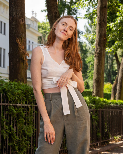 A fair-skinned person with long reddish-brown hair and freckles is standing outdoors with a slight smile. They are wearing a pure white linen cropped wrap top tied at the side with a wide cream-colored ribbon, and stone green pants. The background includes trees, a black metal fence, and a light-colored building.