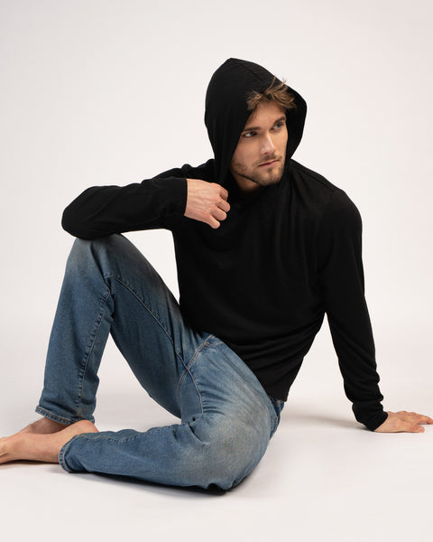 A barefoot man sits on the floor wearing blue jeans and a black long-sleeved hooded shirt with the hood up. He is looking off to the right with one hand resting near his collar and the other on the floor.