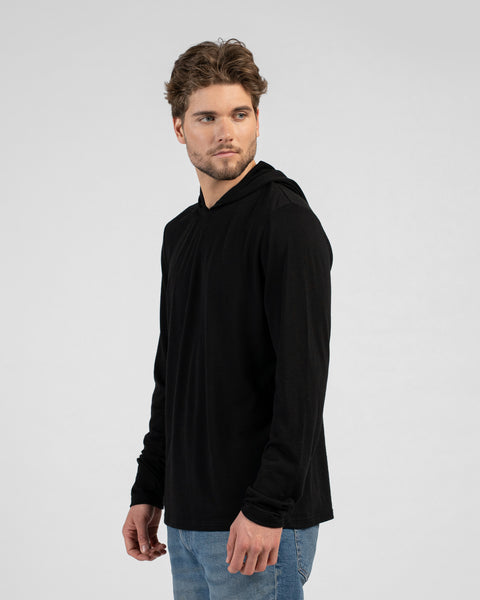 A man stands looking back over his right shoulder. He is wearing a black long-sleeved hooded shirt with the hood resting down and blue jeans against a light grey background.
