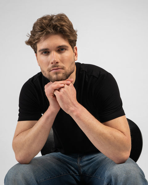 A studio portrait of a man leaning forward in a chair with his chin resting on his clasped hands. He is wearing a black short-sleeved t-shirt and blue jeans, looking directly at the camera.