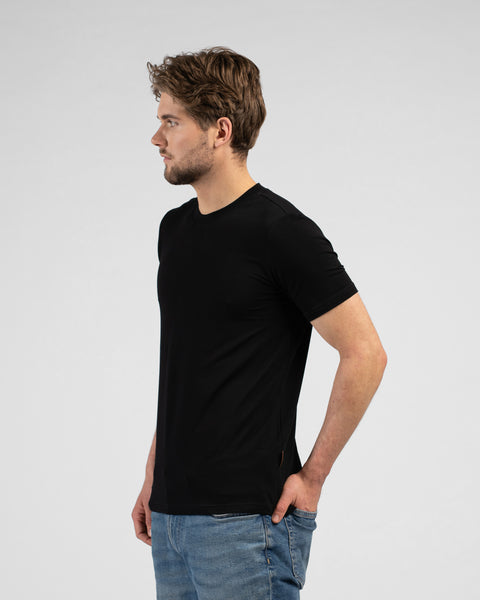 A side profile shot of a man wearing a black short-sleeved t-shirt and blue jeans, facing to the left. He has his right hand resting casually in his front pocket against a light grey background.