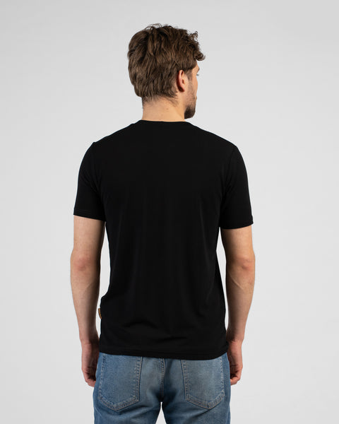 A studio shot showing the back of a man wearing a solid black short-sleeved t-shirt and blue jeans. He stands with his arms relaxed by his sides against a plain light grey background.