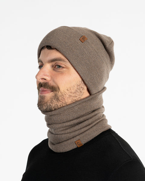 A man with a light brown beard is shown from the chest up, looking to the left of the frame. He is wearing a brown knitted beanie and a matching neck gaiter. Both items of clothing have a small brown leather patch with a menique logo on them. The background is a plain white.