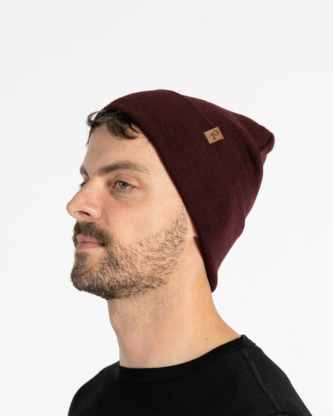 A person with light skin, short brown hair, and a beard is shown in profile against a white background. They are wearing a royal cherry knit beanie with a small brown tag on the side. They are also wearing a black t-shirt.