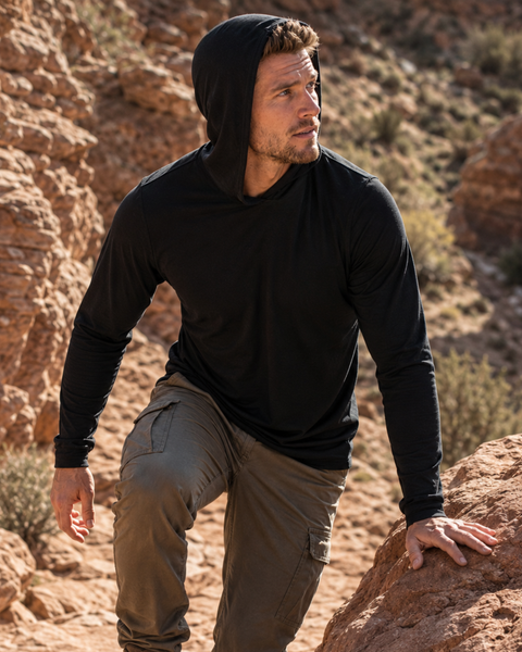 An outdoor shot of a man hiking along a rugged, sunlit rocky trail. He is looking off to the left, wearing a black long-sleeved hooded shirt with the hood down and olive green cargo pants.