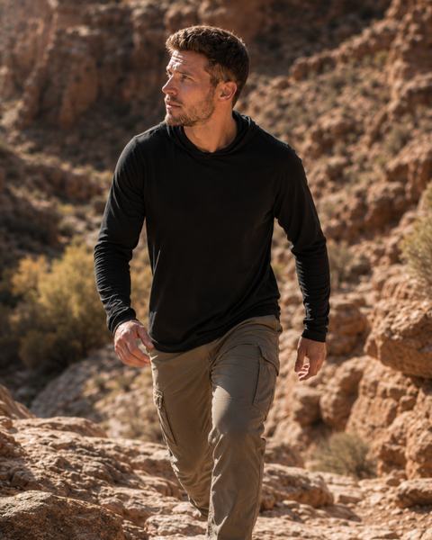 An outdoor shot of a man stepping up onto a large rock in a sunlit canyon setting. He is looking off to the right, wearing a black long-sleeved hooded shirt with the hood pulled up and olive green cargo pants.